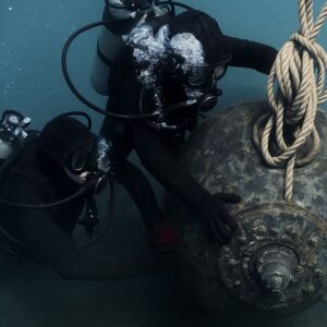 Due palombari della Marina Militare fissano sott’acqua una mina navale utilizzando funi, immersi in un ambiente marino con attrezzature da immersione e bolle d’aria visibili.