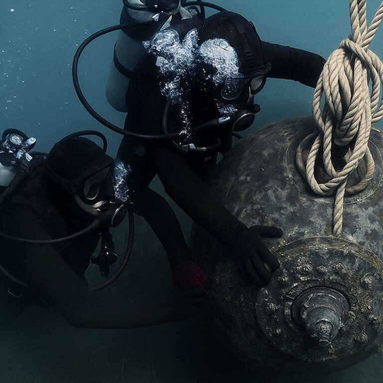 Due palombari della Marina Militare fissano sott’acqua una mina navale utilizzando funi, immersi in un ambiente marino con attrezzature da immersione e bolle d’aria visibili.